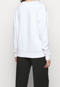 Sudadera blanca de manga larga, con cuello redondo y puños acanalados. Textura suave y corte holgado, mostrada desde la parte de atrás.