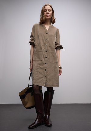 Langærmet brun fløjlskjole med V-hals, knapper foran og to forlommer. Kombineret med knæhøje bordeaux støvler og en tanfarvet taske.