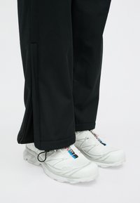 Λευκά αθλητικά παπούτσια με υφασμάτινες σόλες και λευκές κορδόνια σε αντίθεση, σε συνδυασμό με μαύρα παντελόνια που έχουν φερμουάρ στο τελείωμα.