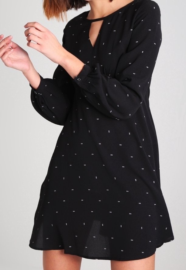 Femme portant une robe noire à manches longues avec de petits motifs en forme de flèche blanche et un décolleté en forme de trou de serrure, ajustant ses manches.