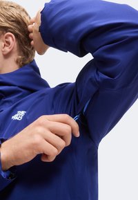 Personne portant une veste bleue déboutonnant la fermeture éclair de ventilation sous le bras côté gauche, montrant une partie de son visage et de ses cheveux.