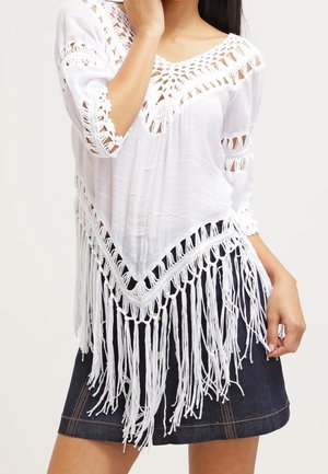 Mujer que lleva una blusa blanca holgada con detalles de crochet y flecos largos combinada con una falda vaquera oscura.