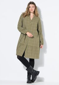 Grünes kariertes Kleid mit langen Ärmeln und V-Ausschnitt, ausgestattet mit einem gestuften Saum. Getragen mit schwarzen Leggings und klobigen schwarzen Stiefeln.