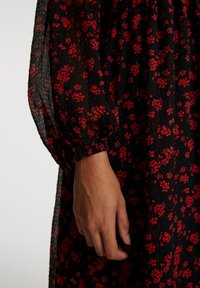 Schwarzes Kleid aus einem strukturierten Stoff mit einem roten Blumenmuster. Puffärmel mit elastischen Bündchen, die einen Teil der Hand zeigen.