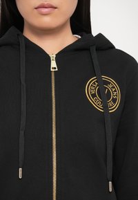 Black zip-up hoodie made of soft fabric, featuring a golden embroidered logo on the chest, and black drawstrings with metal tips.