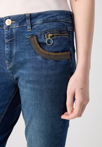 Jean en denim bleu foncé avec une poche zippée et un accent tissé. Dispose d'un détail anneau en métal et de coutures orange. Texture lisse.