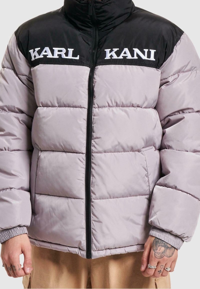 Doudoune gris clair avec épaules noires, fermeture éclair noire et logo blanc « KARL KANI » sur la poitrine, portée avec des mains tatouées visibles.