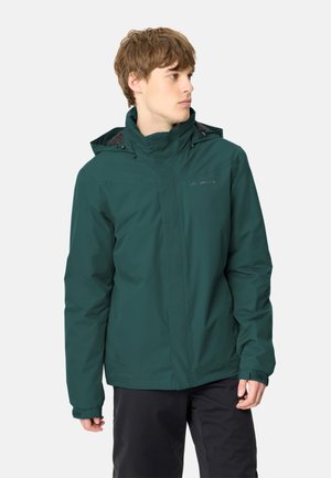Jeune homme portant une veste à capuche vert foncé et un pantalon noir, regardant vers sa gauche sur un fond clair uni.