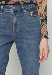 Jeans de ganga azuis com uma cintura alta, apresentando um bolso com fecho prata, costura em contraste e uma textura suave. Um botão em relevo é visível.