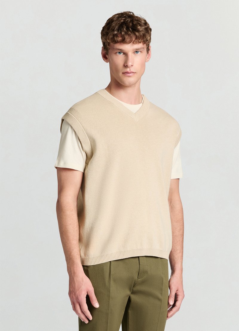 Veste beige tricotée avec un col en V et des manches courtes, portée sur une chemise claire. Texture lisse avec des accents côtelés au niveau du col et de l'ourlet.