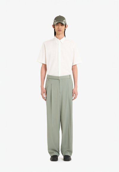 Chemise à manches courtes à rayures claires, pantalon plissé vert menthe, et casquette olive avec lettres blanches. Des chaussures noires complètent l'ensemble.