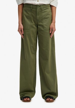 Persona che indossa pantaloni verde oliva a gamba larga con tasche applicate davanti, abbinati a sandali neri aperti e una camicia bianca.