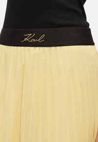 Jupe plissée jaune avec une ceinture noire portant la signature dorée "Karl". Le tissu semble léger et texturé.
