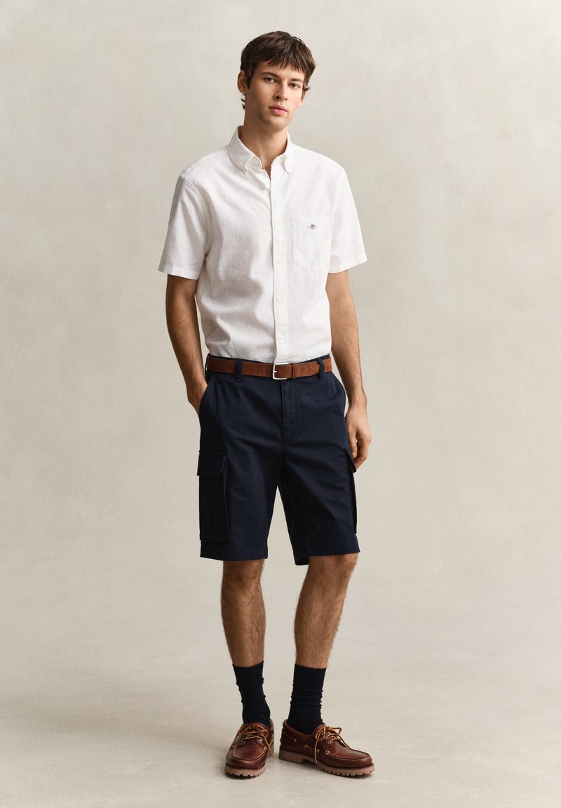 Giovane uomo in piedi, indossa una camicia bianca a maniche corte con bottoni, pantaloncini cargo blu navy, cintura marrone, calzini neri e scarpe di pelle marroni.