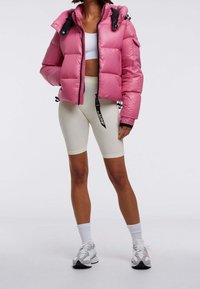Rosa puffjacka med huvtröja, dragkedjestängning och svarta detaljer. Tillsammans med figursydda krämfärgade cykelshorts och grå sportskor.