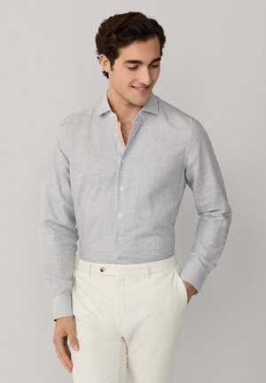 Mann trägt hellblau gestreiftes Hemd mit Knöpfen und weiße Hose, steht mit einer Hand in der Tasche, lächelt und schaut nach unten.