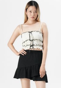 Ung kvinne med rett hår har på seg en hvit rynket crop top med svarte bånd og et svart asymmetrisk volangskjørt, poserer med hånden på hoften.