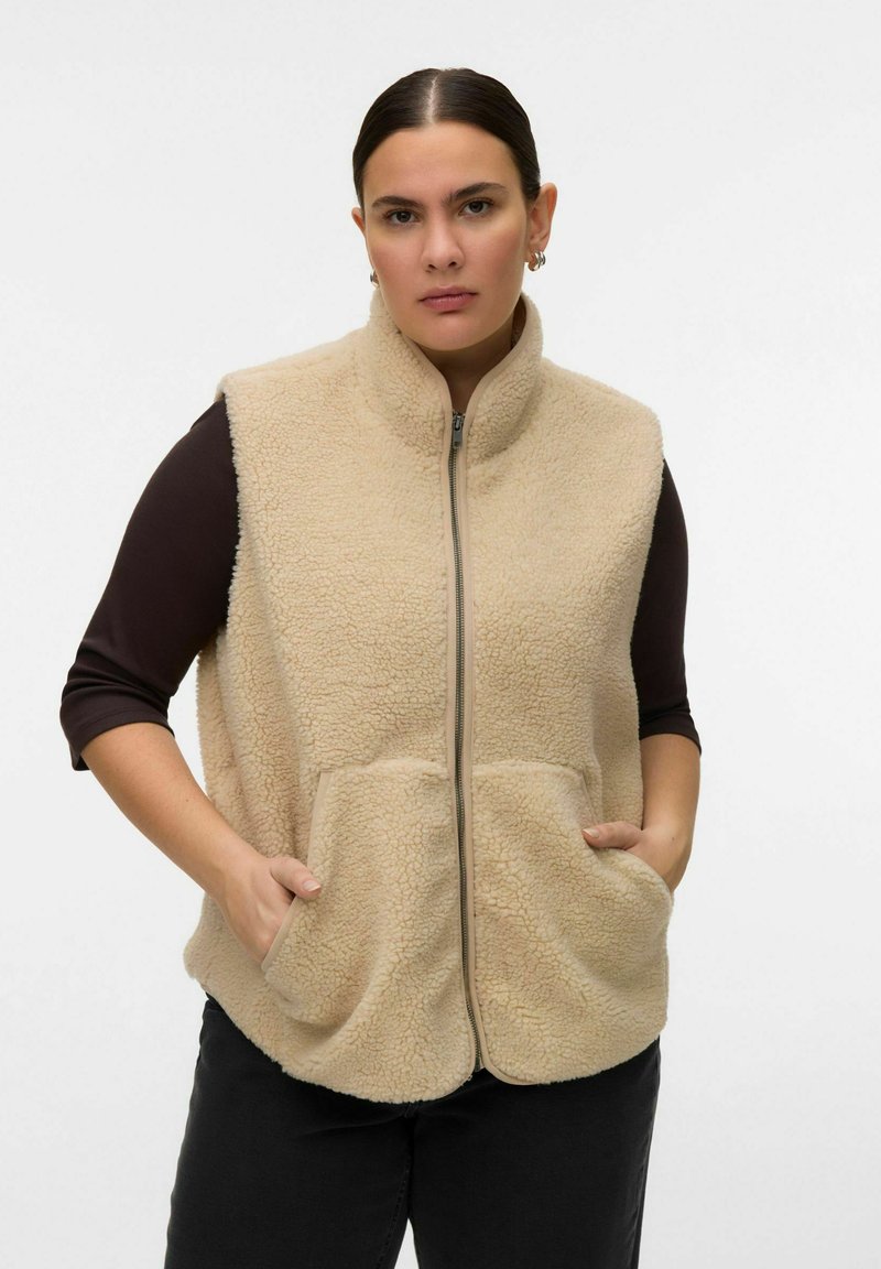 Femme portant un gilet sherpa beige zippé avec poches sur un haut à manches longues noir, debout avec les mains dans les poches du gilet, fond neutre.