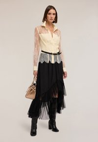 Geel blouse met kanten mouwen, zwarte rok met ruches en doorschijnende stof, zwarte laarzen tot aan de knie, en een kleine beige handtas.