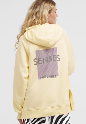 Blonde woman wearing light yellow hoodie with purple square and black text "SENSES" and "THE LABEL" on back, paired with black and white zebra pants.