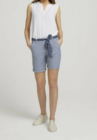 Blusa blanca sin mangas con escote en V, combinada con pantalones cortos enrollados azul claro que cuentan con un cinturón navy estampado, llevada con zapatillas crema.