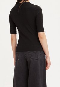 Žena v upnutém černém krátkém rukávu a volných tmavě šedých džínech, zobrazená ze zadního pohledu na jednobarevném světlém pozadí.