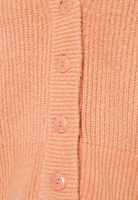 Ljusorange virkad cardigan med ribbat mönster och sex blanka knappar i matchande färg längs fram.