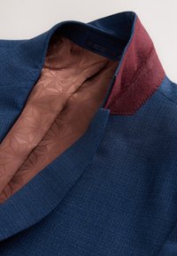 Blazer bleu marine en tissu texturé, avec un col en feutre bordeaux et une doublure marron à motifs. Présente un design moderne et ajusté.