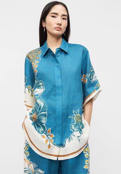 Νεαρή γυναίκα με μακριά σκούρα μαλλιά φοράει ένα μπλε πουκάμισο με λουλούδια, κουμπωτό, με φαρδιά μανίκια και ταυτόσημο παντελόνι μπροστά σε απλό φόντο.