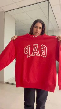 Jeune femme tenant un sweat rouge GAP avec des lettres blanches dans une pièce intérieure avec des murs miroirs et un plafond carrelé.