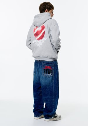Gray hoodie with red and white graphic apple design on back; paired with loose blue denim jeans featuring a logo patch on the pocket.
