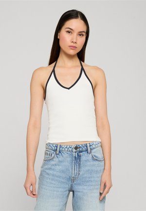 Jeune femme aux longs cheveux noirs portant un débardeur blanc à liseré noir et un jean taille haute bleu clair, debout devant un fond uni.