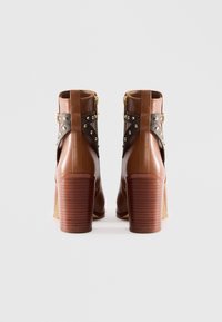 Brown leather ankle boots with stacked heels, black studded straps, and gold side buckles, shown from the back on a white background.