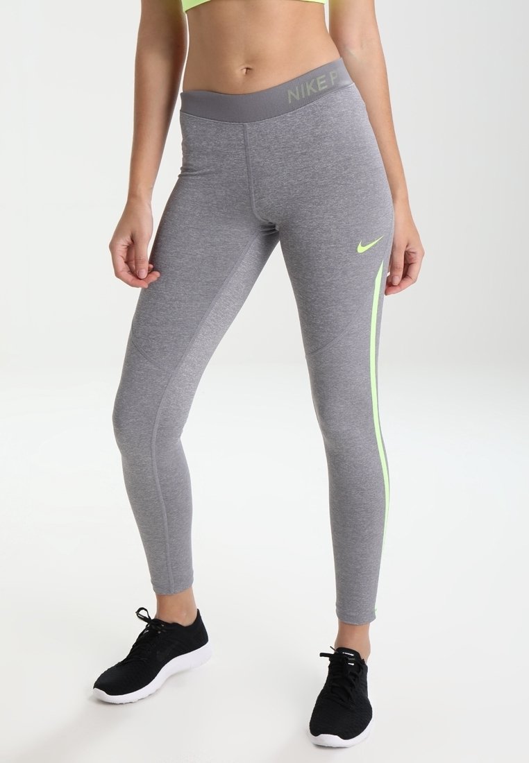 Mujer vistiendo leggings grises de Nike con acentos neón y zapatillas negras, de pie contra un fondo blanco liso.