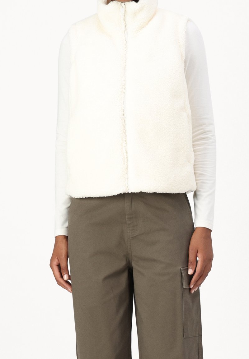 Weißer, flauschiger Fleece-Weste mit hohem Kragen, Frontreißverschluss und ohne Ärmel, kombiniert mit olivgrünen Cargo-Hosen. Glatte Textur und minimales Design.