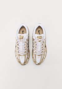 Białe i złote buty sportowe Nike z siateczkową cholewką, zaokrąglonym noskiem i wyraźnym systemem sznurowania. Złote akcenty i czarne logo na języku.