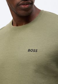 Κοντινό πλάνο ενός άνδρα που φοράει μια υφασμάτινη πουκάμισο ελιάς με τη λέξη "BOSS" κεντημένη στο στήθος.