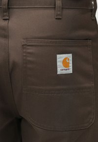 Καφέ παντελόνι εργασίας με πίσω τσέπη που έχει ένα λευκό σημείο με πορτοκαλί λογότυπο. Το ύφασμα είναι ανθεκτικό με υφή.