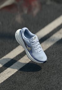 Svijetloplava Nike trkačka cipela s bijelim potplatom u zraku iznad asfaltirane ceste s bijelom linijom.