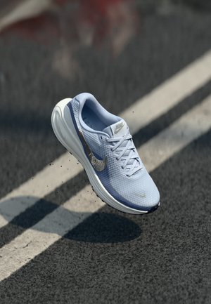 Svijetloplava Nike trkačka cipela s bijelim potplatom u zraku iznad asfaltirane ceste s bijelom linijom.