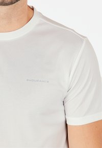 Hvid kortærmet atletisk t-shirt lavet af letvægtsstof, der har et diskret tekstureret mønster og et lille gråt logo på brystet.