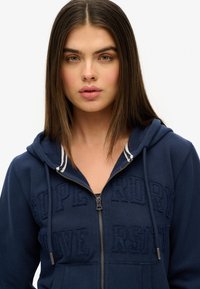 Sweat à fermeture éclair bleu marine avec des accents à rayures blanches, présentant des lettres en relief sur le devant et une capuche avec cordon de serrage. Tissu en mélange de coton.