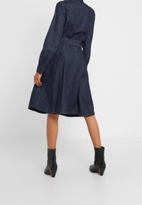 Denimklänning, mörkblå, långa ärmar, bälte i midjan, utsvängd kjol, enfingerknapp vid ärmslut och svarta klackboots som når anklarna.