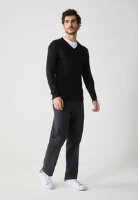 Uomo in piedi che indossa un maglione nero a V, pantaloni grigio scuro e sneakers bianche, contro uno sfondo chiaro e semplice.