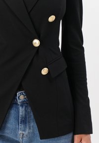 Zwarte blazer met een dubbelzijdig ontwerp, gouden knopen met sterrenreliëf, op maat gemaakte pasvorm en een gestructureerde stof. Draag over spijkerbroeken.