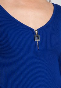 Nahaufnahme eines blauen V-Ausschnitt-Oberteils mit Rippstruktur und einem silbernen quadratischen Reißverschluss-Zipper am Halsausschnitt.