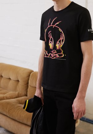 Personne portant un t-shirt noir avec un motif coloré de Tweety Bird, tenant une veste noire et une casquette, debout à l'intérieur près d'un canapé beige.