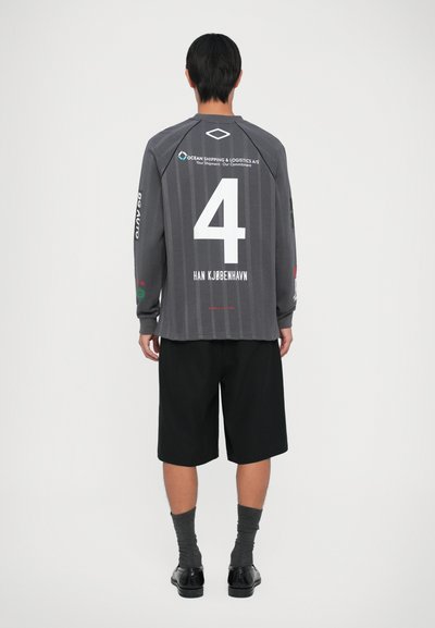 Pessoa de pé de costas, vestindo uma camisola cinza de mangas longas com o número 4, calções pretos, meias cinzentas e sapatos pretos, num fundo branco.