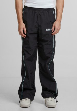 Person trägt schwarze Jogginghose mit hellblauen Paspeln und weiße Turnschuhe, steht vor einem schlichten grauen Hintergrund.