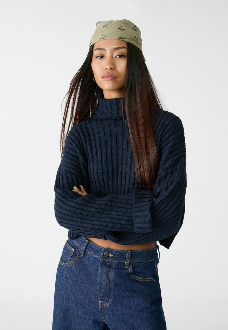 Navy ribbel gebreide trui met een hoge kraag en brede mouwen, gecombineerd met blauwe high-waisted jeans. Groene met patroon hoofddoek.